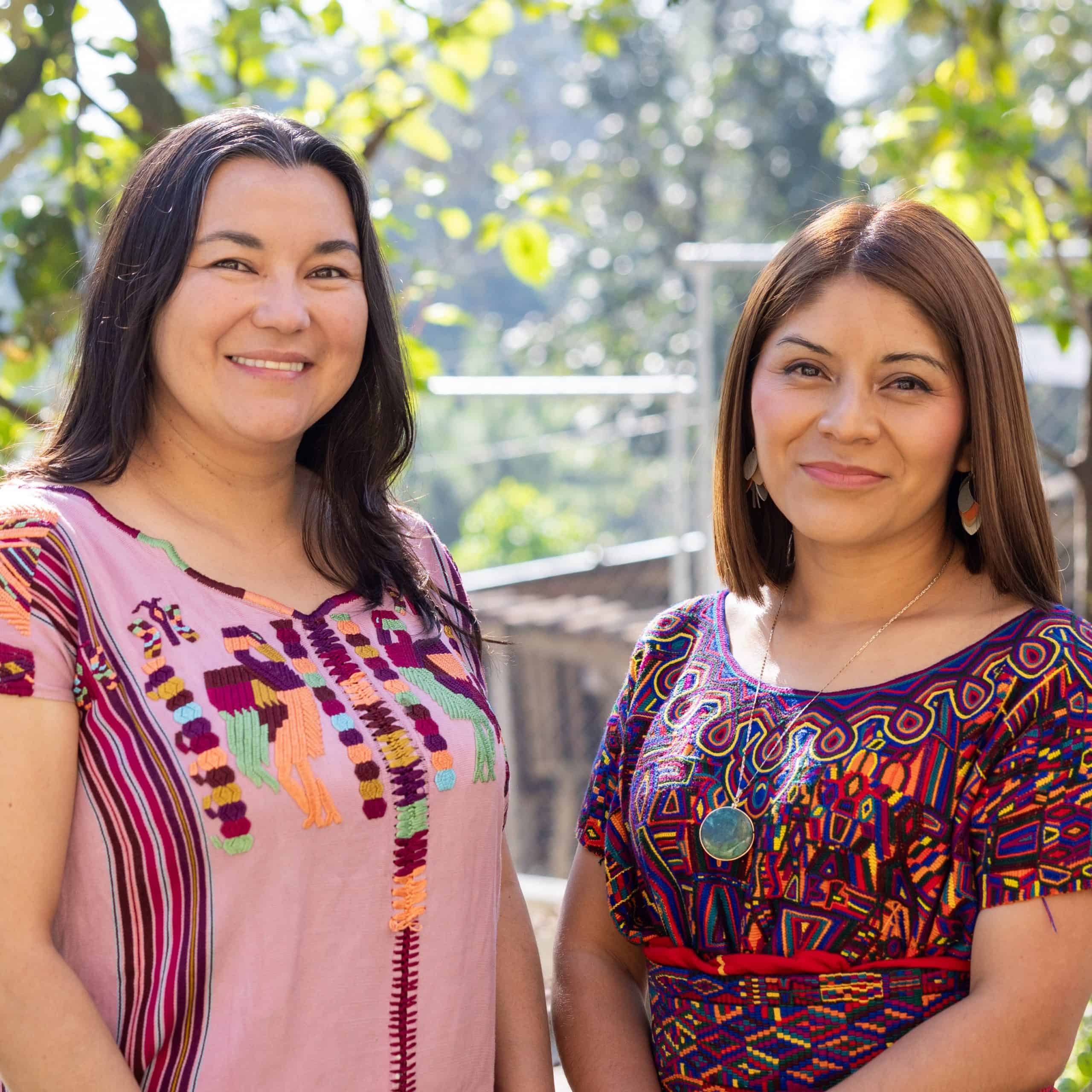 Two indigenous women smiling outdoors, wearing colorful traditional clothing.