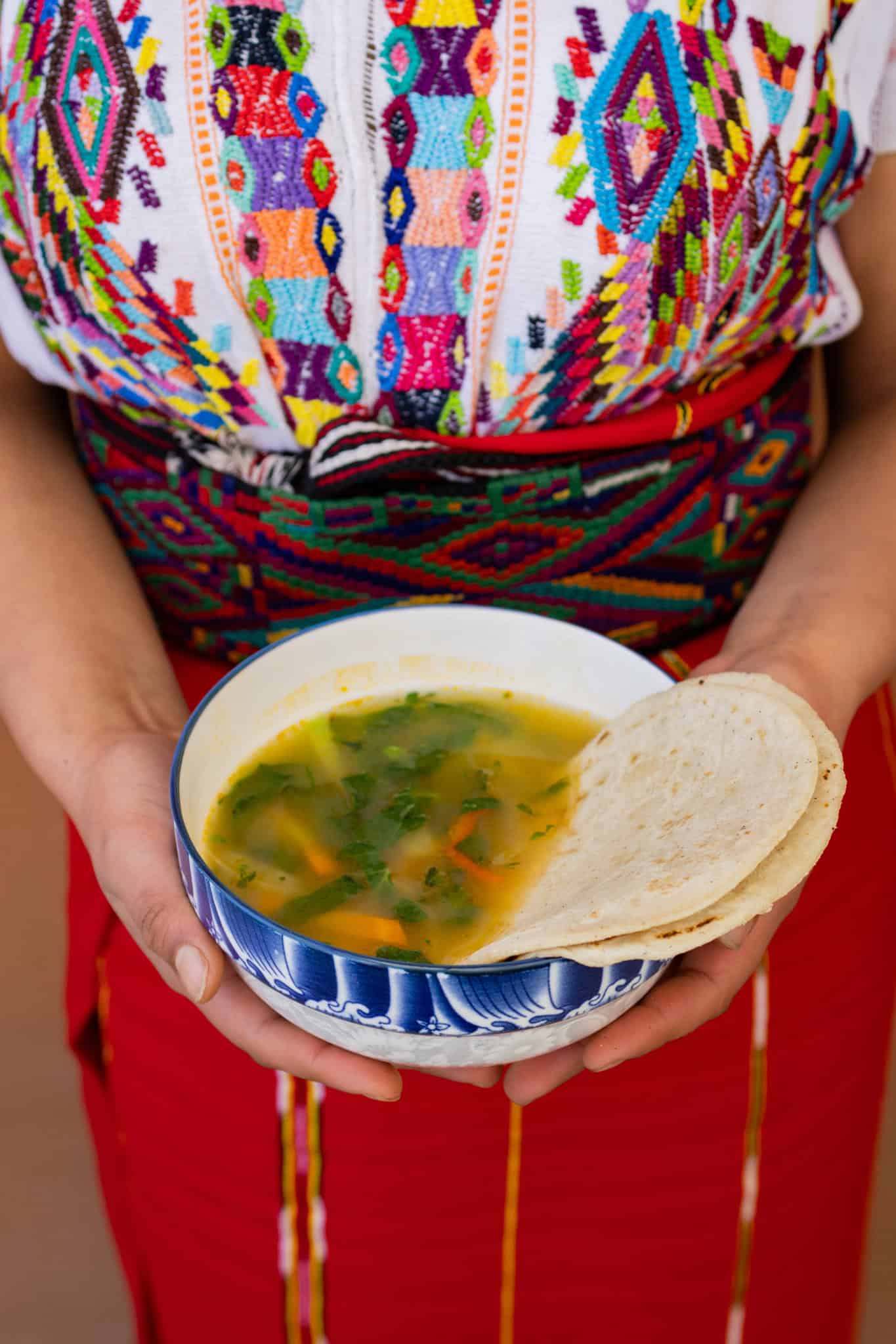 Traditional Ixil woman with colorful embroidered clothing holding soup and bread.
