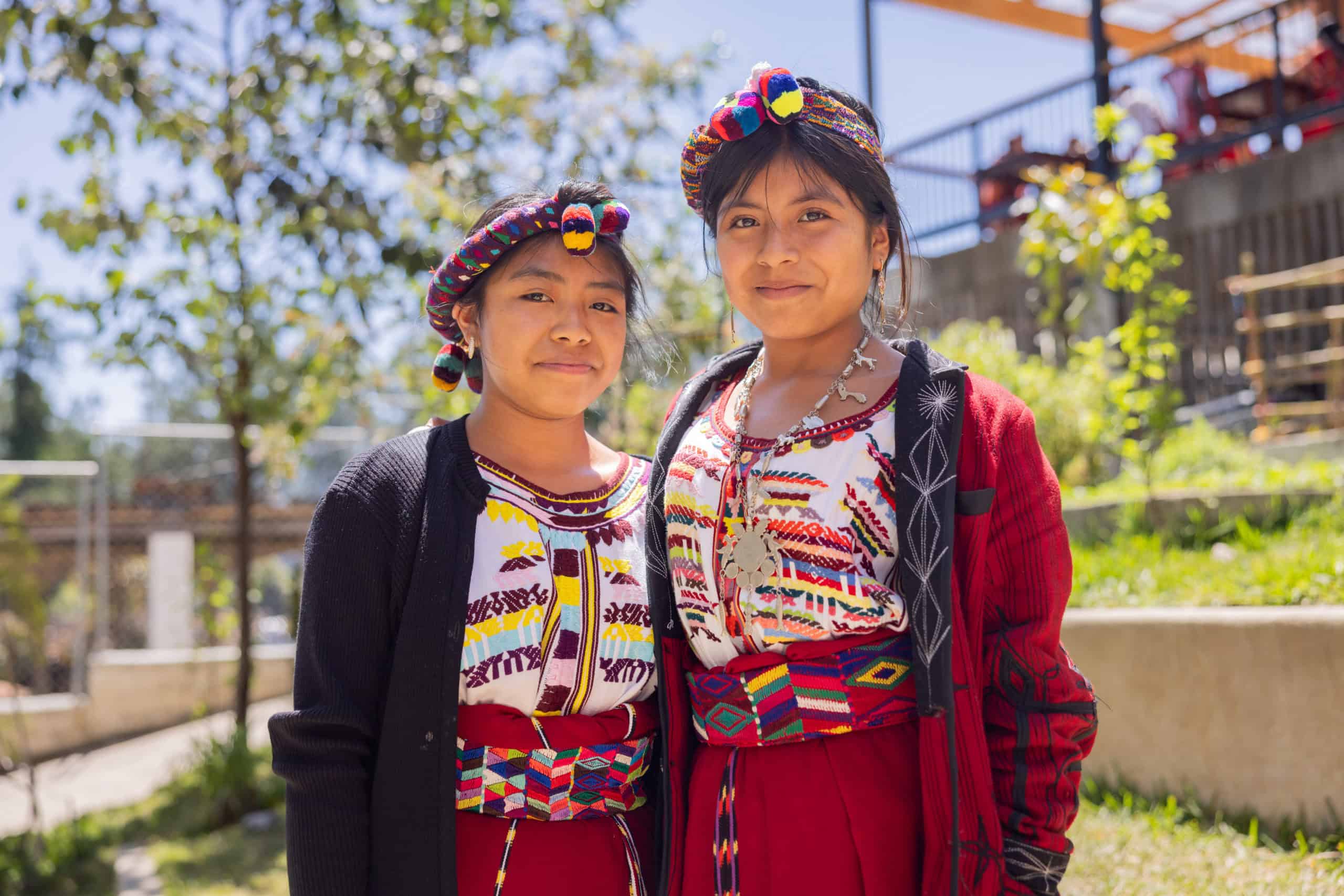 Two Ixil girls in traditional attire smiling outdoors in a community setting.