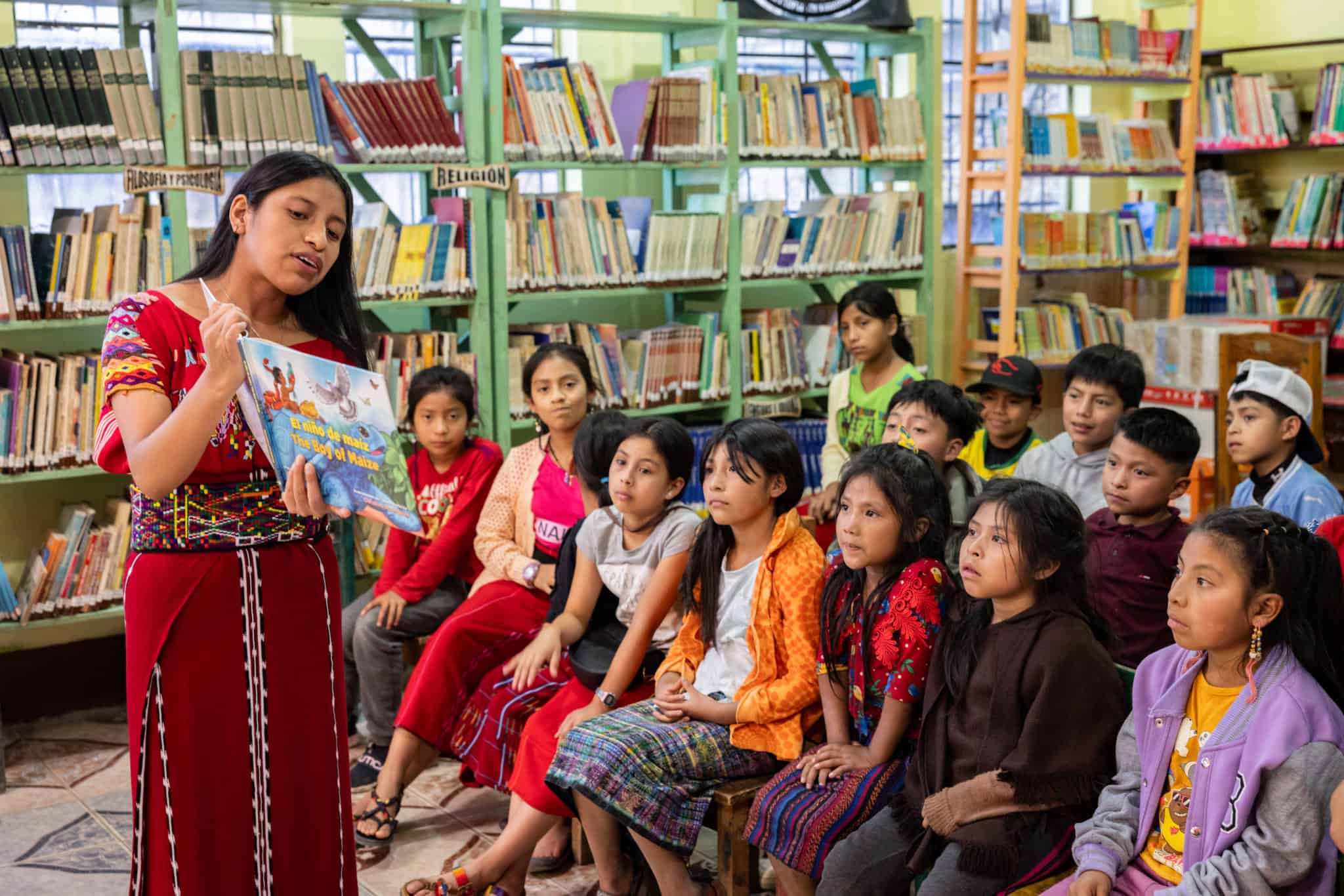 Woman reading to children in a library with bookshelves in the background.