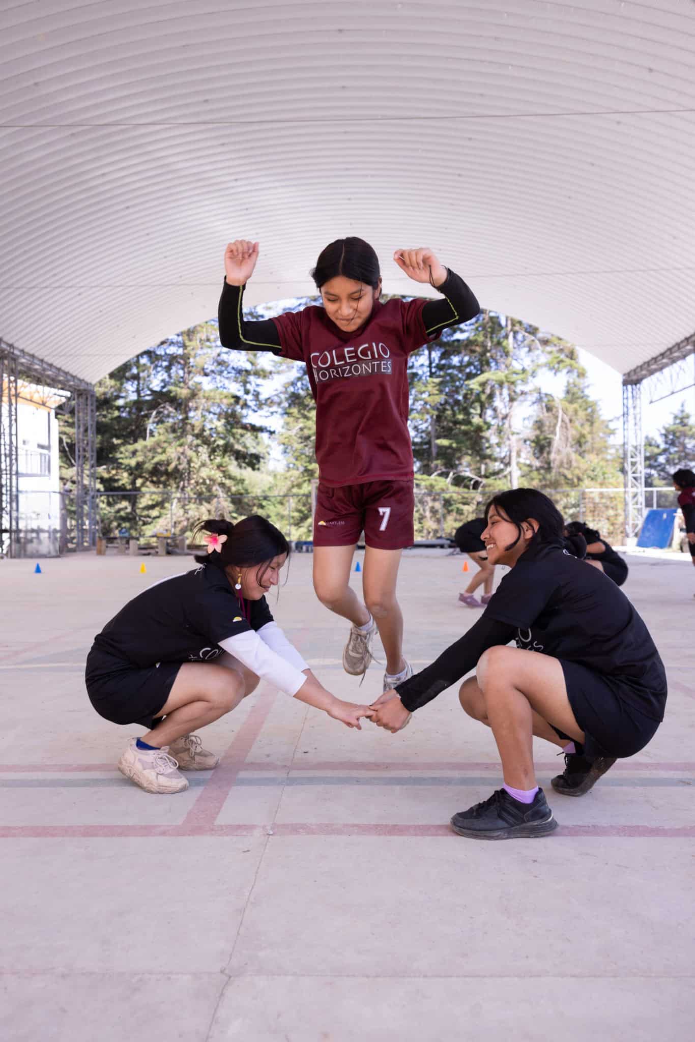 Young girl jumping with support from two peers under a covered outdoor area.