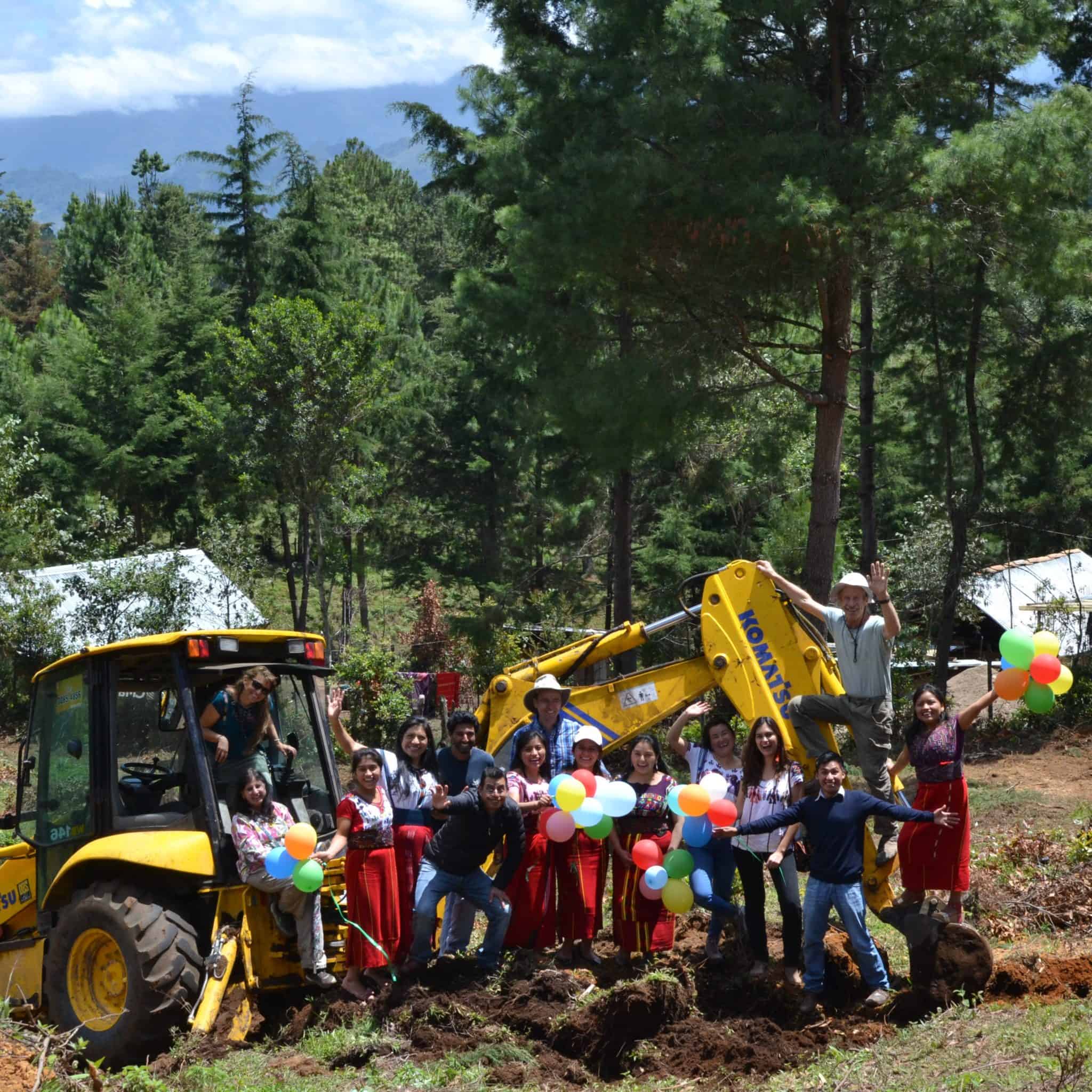 Group of people with balloons and a yellow excavator in a forested area.